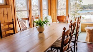 Una mesa de comedor de madera con sillas, un florero con una planta y amplias ventanas con vistas a un lago y árboles.