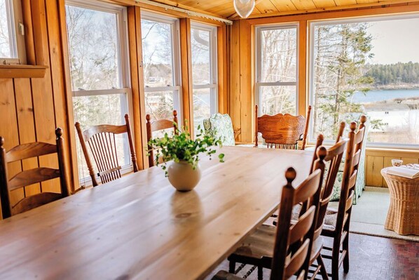 Una mesa de comedor de madera con sillas, un florero con una planta y amplias ventanas con vistas a un lago y árboles.
