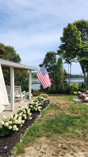Une terrasse couverte, aménagée avec du mobilier d’extérieur, un drapeau et un jardin où poussent des hortensias.