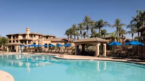 Una piscina con sillas de descanso y sombrillas, rodeada de palmeras y un edificio con varios balcones.