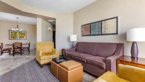 Habitación de hotel con un sofá de cuero marrón, un sillón amarillo, una mesita de centro de madera y una zona de comedor con mesa y sillas.
