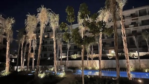 A nighttime view of a hotel with multiple floors, palm trees, and a swimming pool. - Chic 2-bedroom apartment with Private Garden + Pool view At Villette (Cairo Governorate)