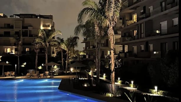 A nighttime view of a hotel pool area with multiple floors of buildings, palm trees, and lounge chairs. - Chic 2-bedroom apartment with Private Garden + Pool view At Villette (Cairo Governorate)