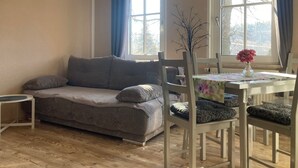A living room with a sofa, a dining table with chairs, a vase with flowers, and a window with curtains. - Holiday Apartment by the Zschopau River (Niederwiesa)