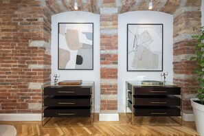 Une pièce avec un mur en briques, deux meubles en bois foncé dotés de poignées dorées, et des œuvres d’art abstrait accrochées au mur.