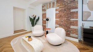 Un salon moderne avec un mur d’accent en briques, deux fauteuils blancs, une table basse ronde, un lampadaire et une plante en pot.