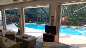 A living room with a pool view, wicker furniture, a television, and a clock on the wall. - Tropical paradise getaway on the Treasure Coast -FL- (Jensen Beach)