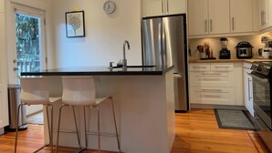 Moderni keittiö, jossa on keskeinen saareke, ruostumattomasta teräksestä valmistetut kodinkoneet, valkoiset kaapit ja riippuvalaisimet.