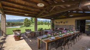 Patio esterno con tavolo di legno apparecchiato per mangiare, circondato da sedie, con vista su una piscina e vegetazione.