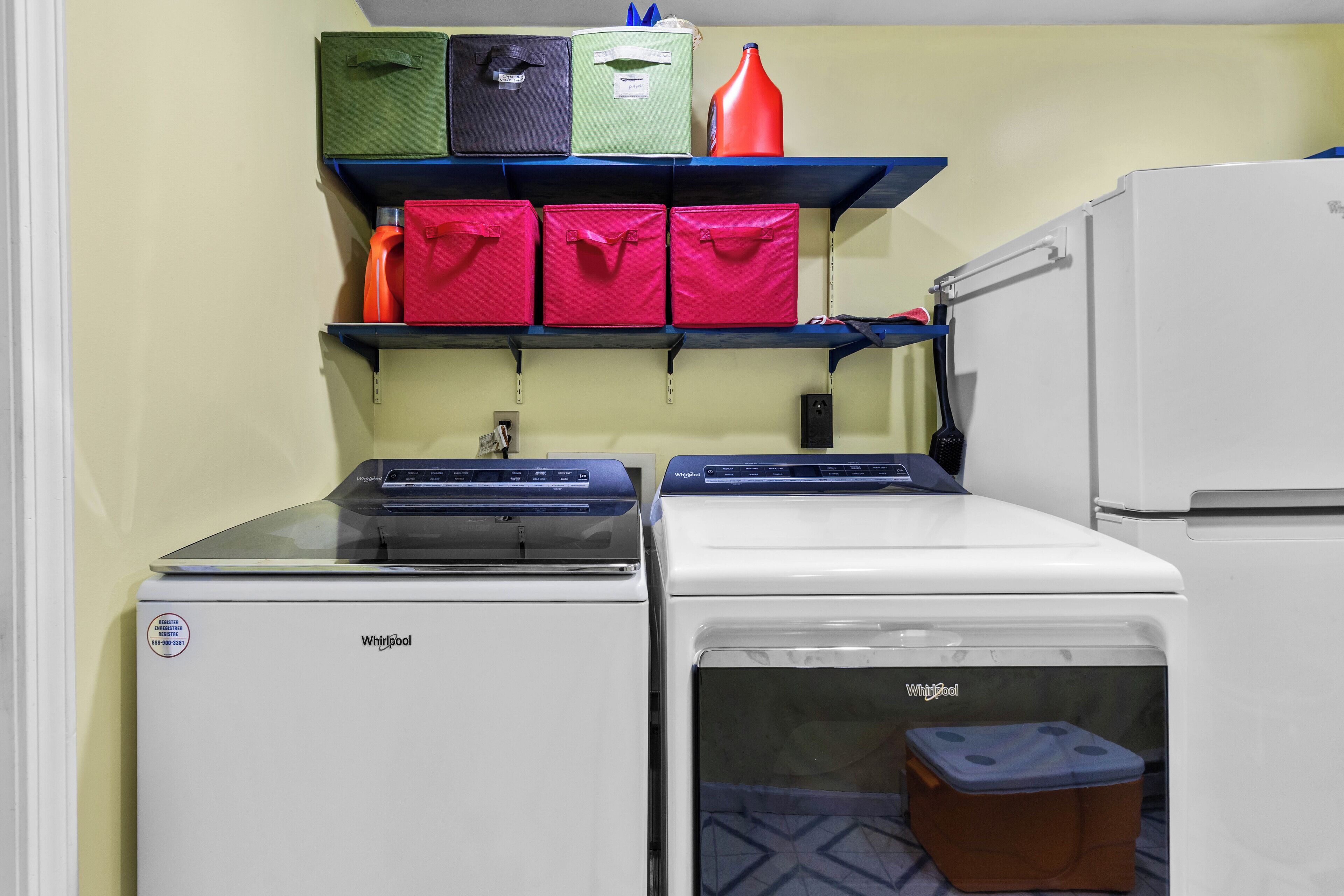 A compact kitchen with two Whirlpool appliances, a shelf with storage boxes, and a small trash bin.