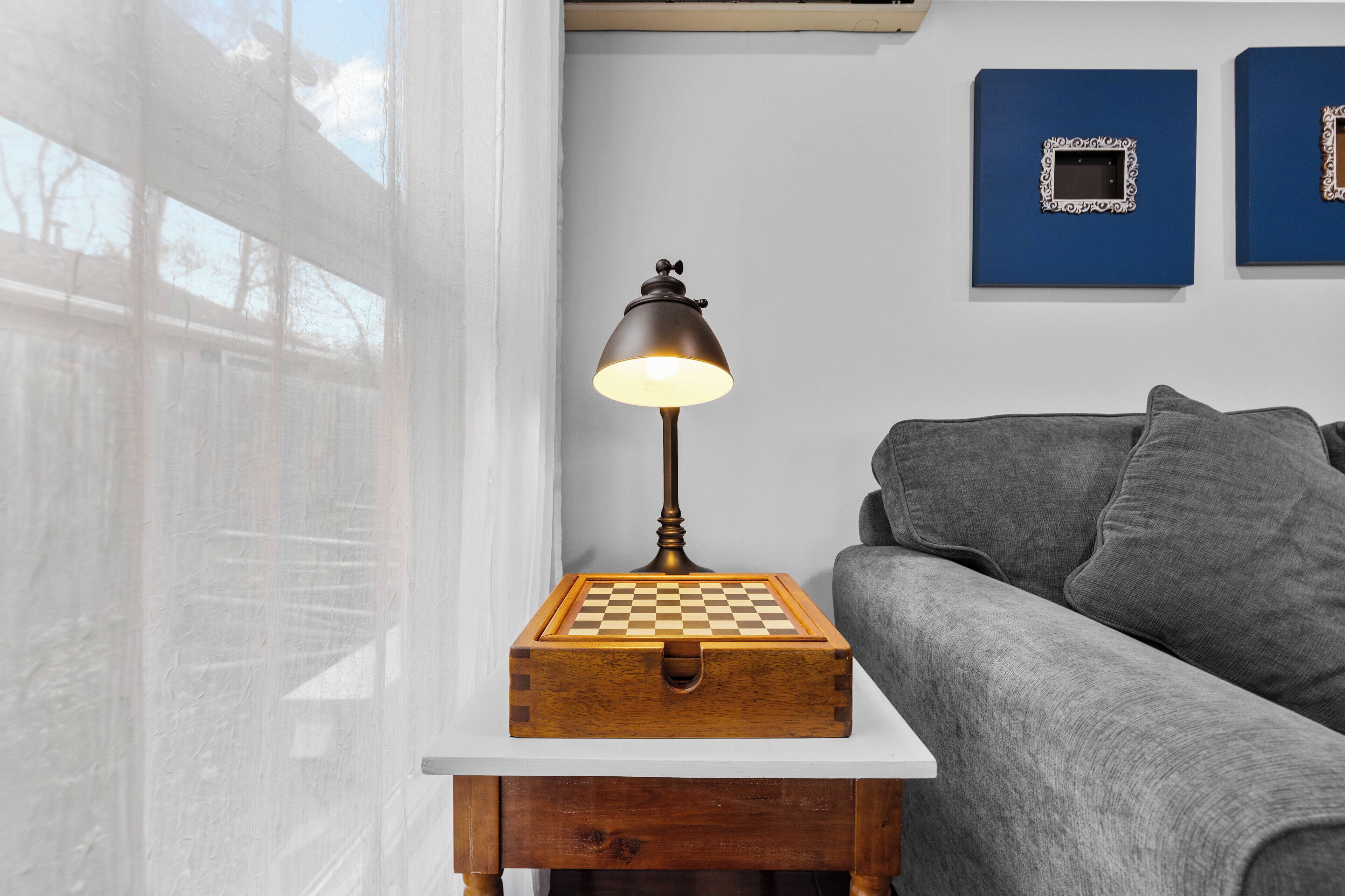 Een houten nachtkastje met een schaakbord, een lamp en een grijze bank in een kamer met een witte muur en een raam.