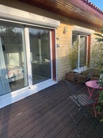 Una terraza de madera con puerta corrediza de vidrio que da a una habitación con cama y baño. Hay plantas en macetas y una pequeña mesa con sillas.