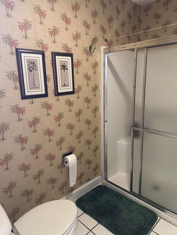 A bathroom with a toilet, a shower with a glass door, and patterned wallpaper. - Common 1 Brunswick Plantation (Calabash)