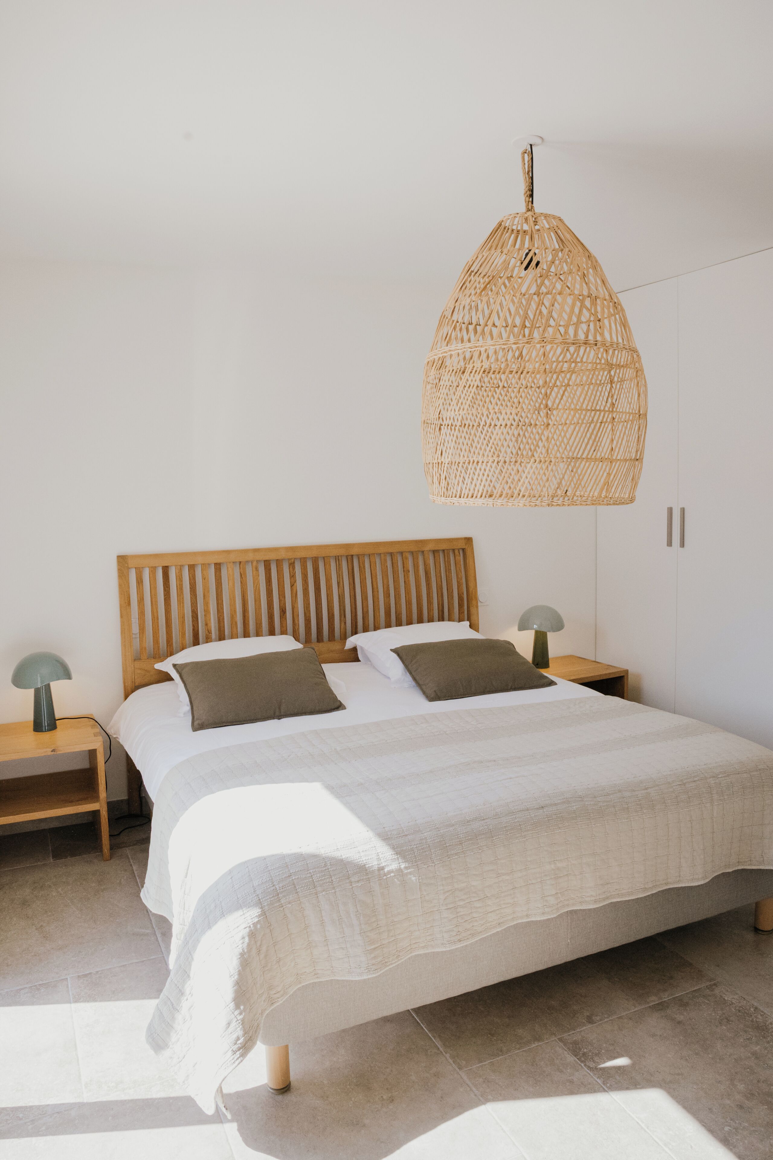 A bedroom with a wooden headboard, a bed with white bedding, a woven hanging light fixture, and a nightstand with a lamp.