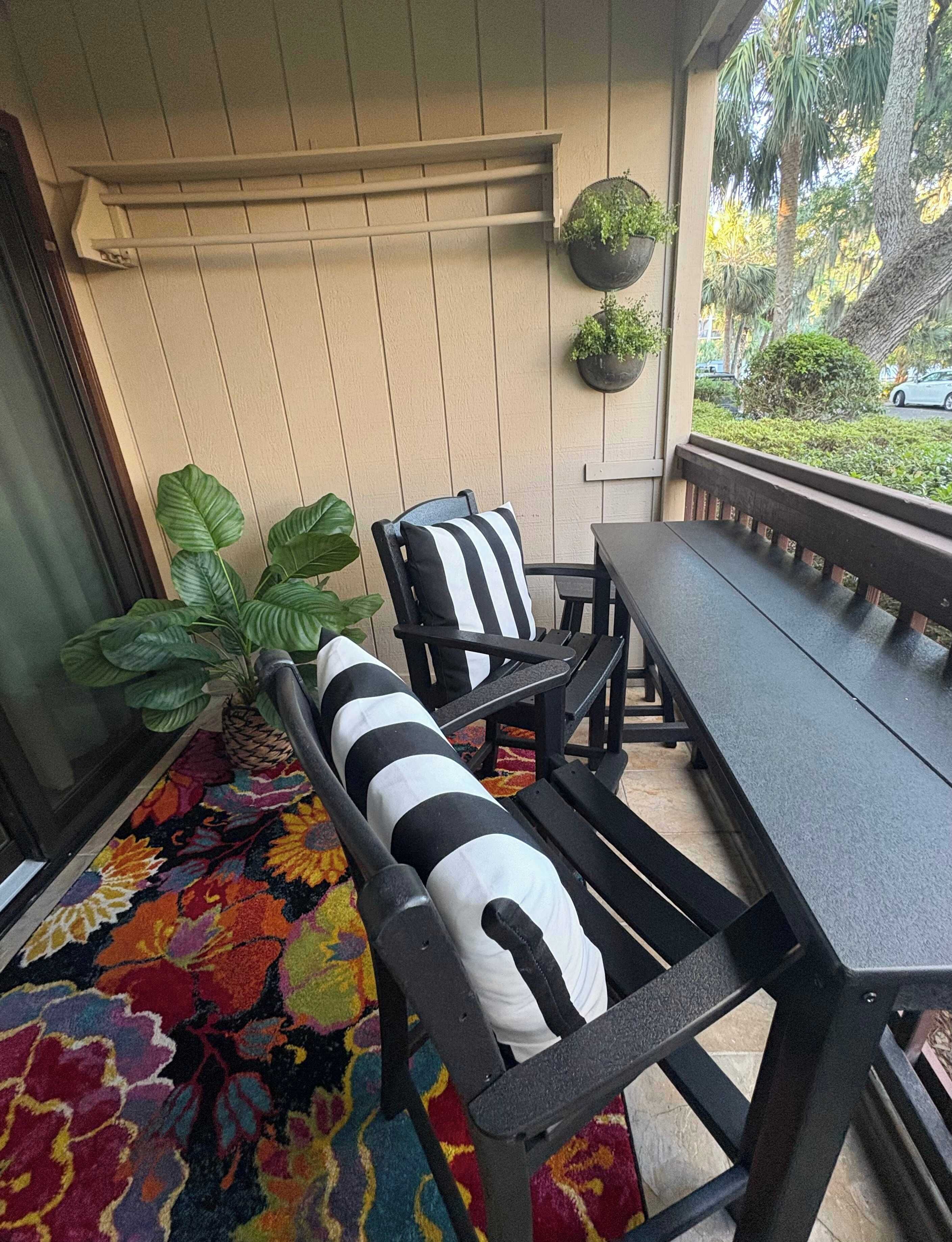 Une terrasse avec une table et des chaises en métal noir, un tapis coloré et une plante en pot.