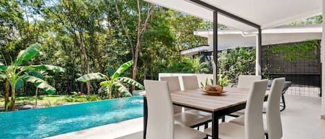 A modern outdoor dining area with a pool, surrounded by lush greenery.