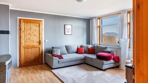 A room with a grey sectional sofa, a wooden door, a window with a view, and a radiator.