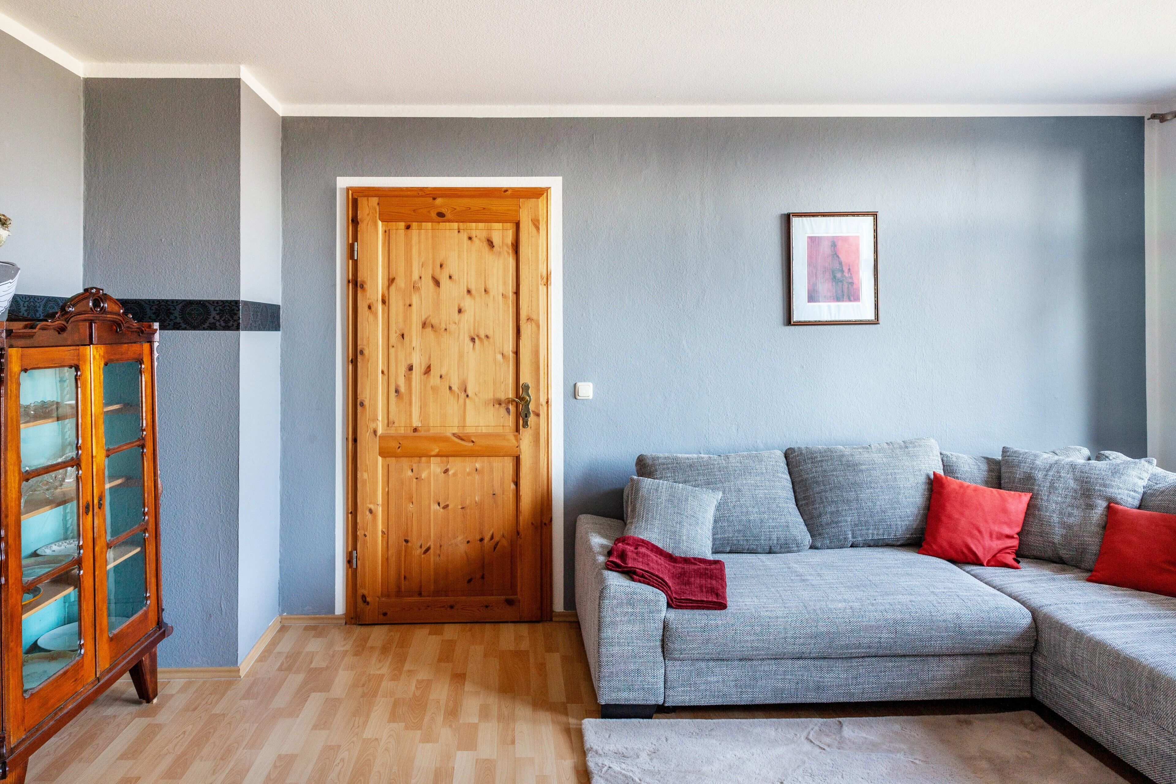 A living room with a wooden door, a grey sofa with red cushions, and a framed picture on the wall.