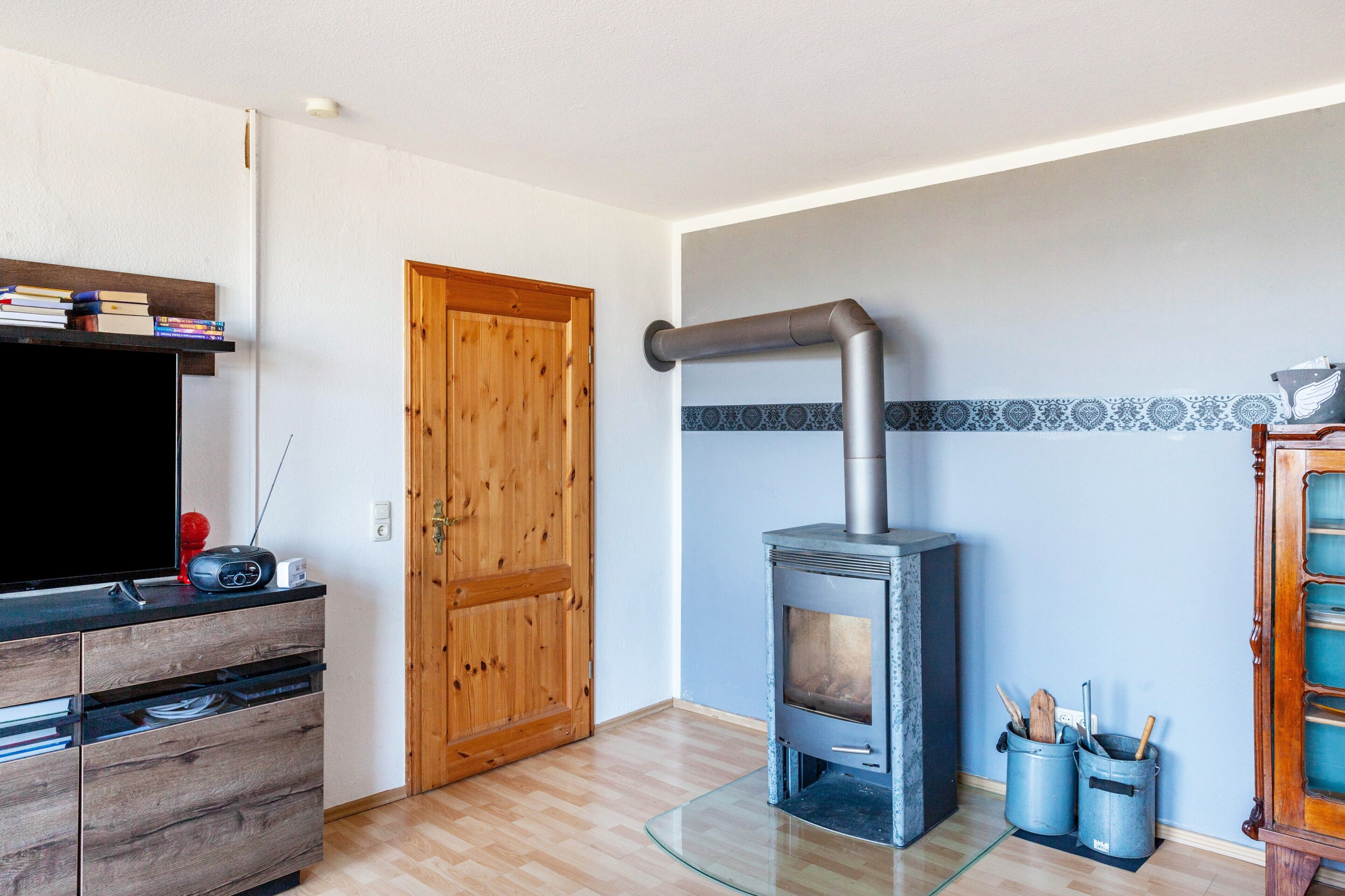Ein Zimmer mit Holztür, eingebautem Kaminofen, einem Glastisch und einer Holzkassette mit Schubladen.
