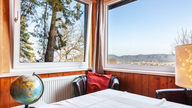 A room with a bed, a globe, a window with a view of trees and hills, and a lamp.