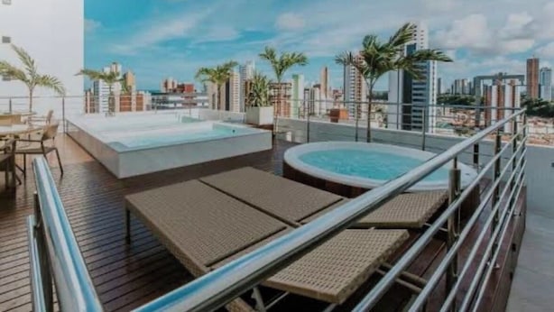 A rooftop pool area with lounge chairs and a hot tub, surrounded by palm trees and city buildings in the background. - Flat Aconchegante em João Pessoa (João Pessoa)