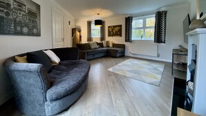 A modern living room with a curved grey sofa, a world map on the wall, and a large window with patterned curtains. - Bracknell / Wokingham Charming Three Bedroom House (Bracknell)