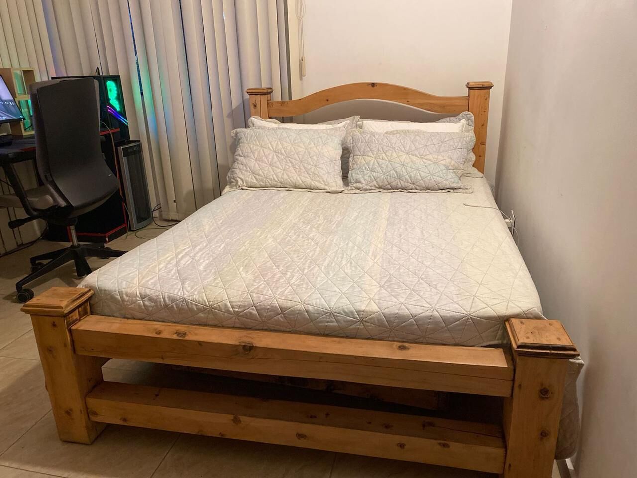 A wooden bed with a quilted mattress and pillows, a wooden headboard, and a desk with a chair in the background.