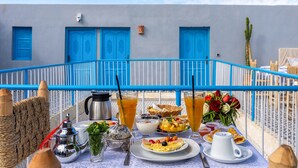 A table set with breakfast items, a metal pitcher, a glass of orange juice, and a vase of flowers on a balcony with blue railings and doors. - Riad Les Secrets De Marrakech (Marrakech)