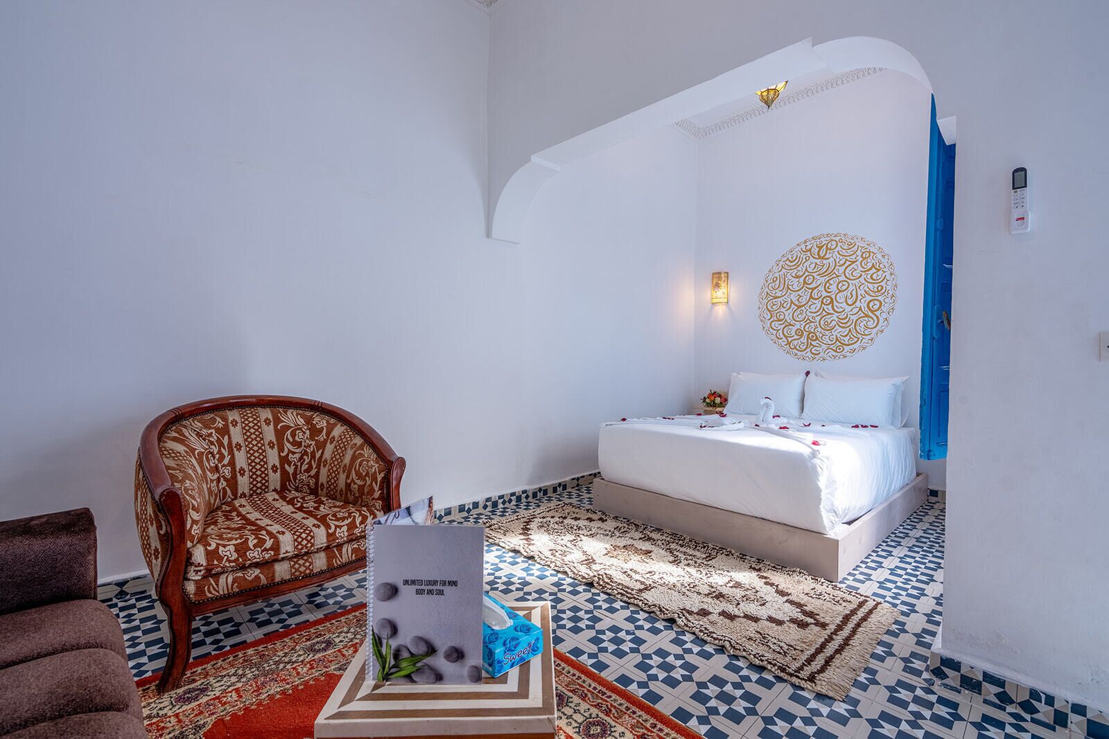 A room with a bed, a chair, a decorative wall piece, and a small table with a sign.