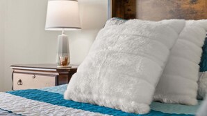 Una cama con colcha azul y blanca, un almohadón mullido blanco, una lámpara de noche y un cabecero de madera.