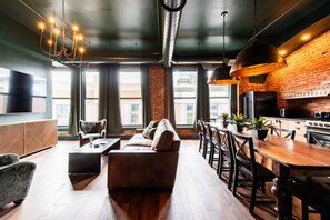 Ein modernes Wohnzimmer mit Sofa, Couchtisch, Esstisch und Stühlen. Es gibt eine Küchenzeile mit Herd und Kühlschrank. Der Raum hat große Fenster und eine Ziegelwand.