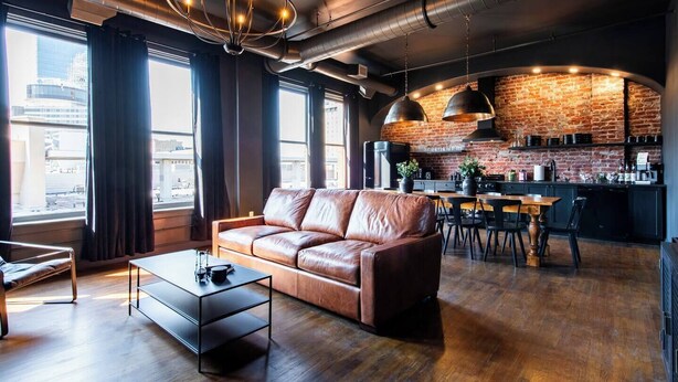 Un soggiorno moderno con divano in pelle, tavolo da pranzo e angolo cottura con pareti in mattoni a vista e illuminazione industriale.