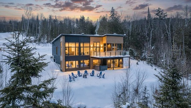 Une maison moderne à deux étages, dotée de grandes fenêtres et d’un balcon, entourée d’une forêt enneigée.
