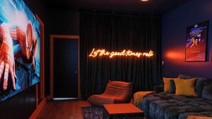 A cozy room with a large screen displaying a movie, a neon sign reading "Let the good times roll," a plush sofa, and a small coffee table. - Mini Golf|Sauna|Hot Tub|Cinema|Large Group|BBQ (Indianapolis)