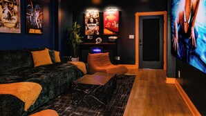 A dark-themed living room with a black sofa, orange cushions, a marble coffee table, and a large flat-screen TV on the right. - Mini Golf|Sauna|Hot Tub|Cinema|Large Group|BBQ (Indianapolis)