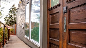 A wooden front door with a keypad lock, a window with a view of a tree and a building, and a sidewalk with a metal railing. - Mini Golf|Sauna|Hot Tub|Cinema|Large Group|BBQ (Indianapolis)