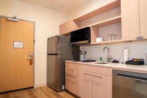 A compact kitchen with a stainless steel refrigerator, a microwave, and a sink with a faucet. There are cabinets above the countertop and a pet-in-room sign on the door.
