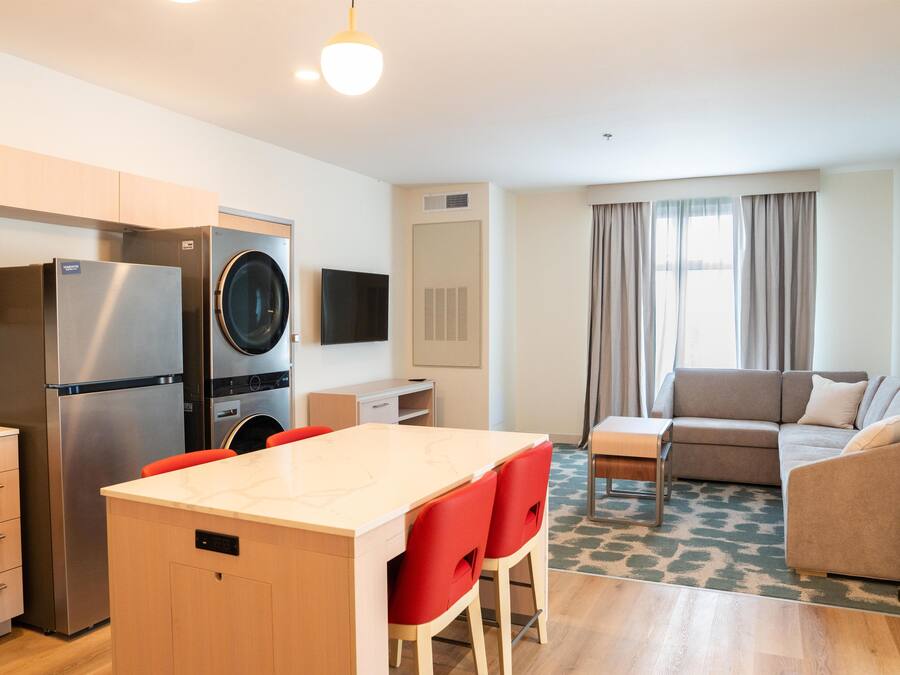 Een compacte hotelkamer met kitchenette, een zithoek met rode stoelen, een bank en een groot raam met gordijnen.