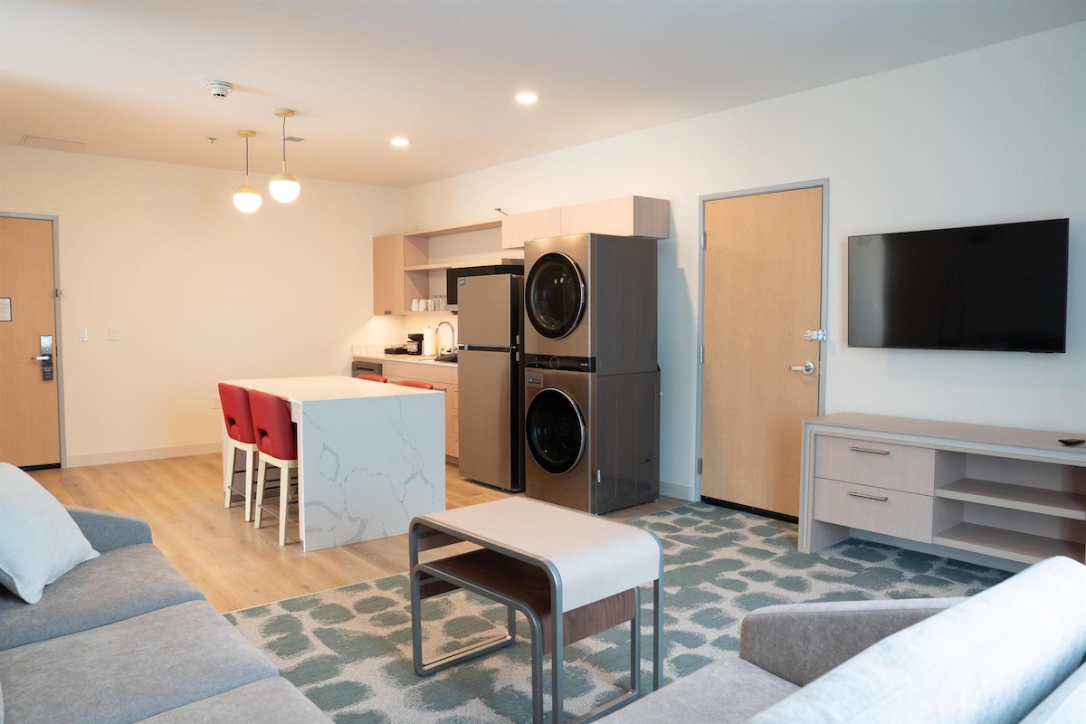 A modern apartment with a kitchenette, a dining area, a living room with a sofa, and a TV mounted on the wall.
