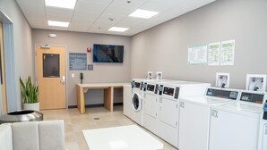 A guest laundry area with a table, chairs, and multiple washing machines. - Homewood Suites By Hilton Corpus Christi Downtown (Corpus Christi)