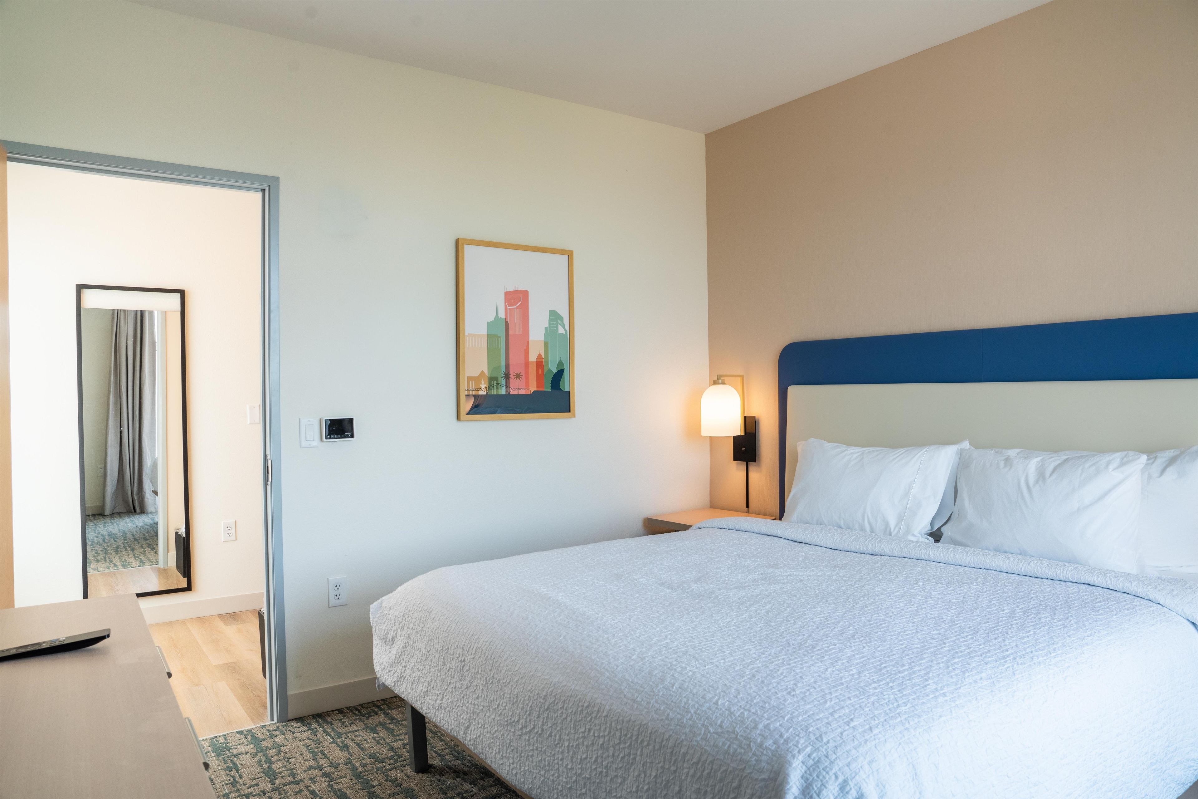 Une chambre d’hôtel avec un lit, une table de chevet, une lampe fixée au mur, un tableau et une porte donnant sur une autre pièce.