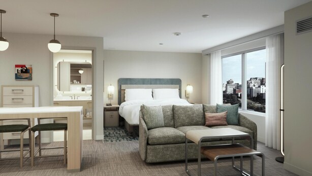 A hotel room with a bed, a sofa, a desk with two chairs, a window with a city view, and a bathroom with a mirror and sink. - Homewood Suites By Hilton Corpus Christi Downtown (Corpus Christi)