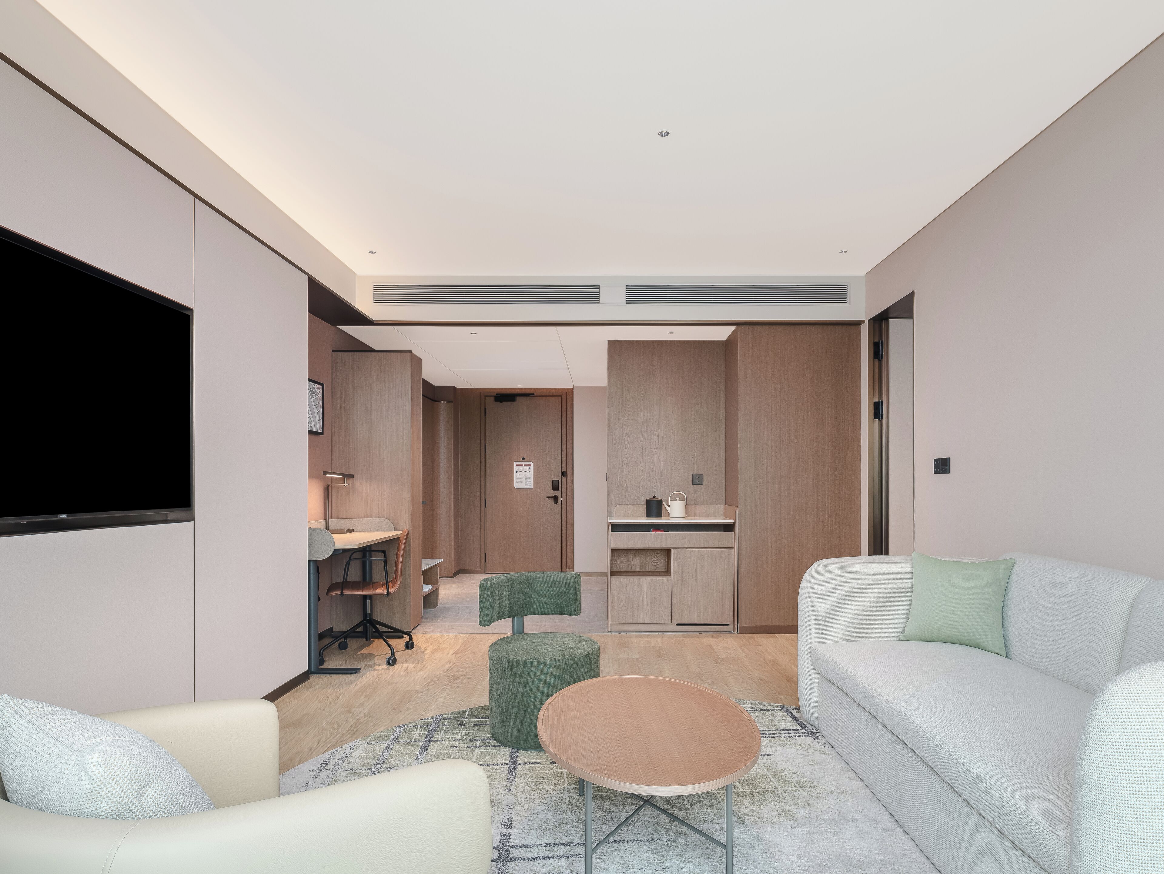 Una habitación de hotel moderna con sofá, una pequeña mesa redonda, un escritorio con silla, kitchenette y televisor montado en la pared.