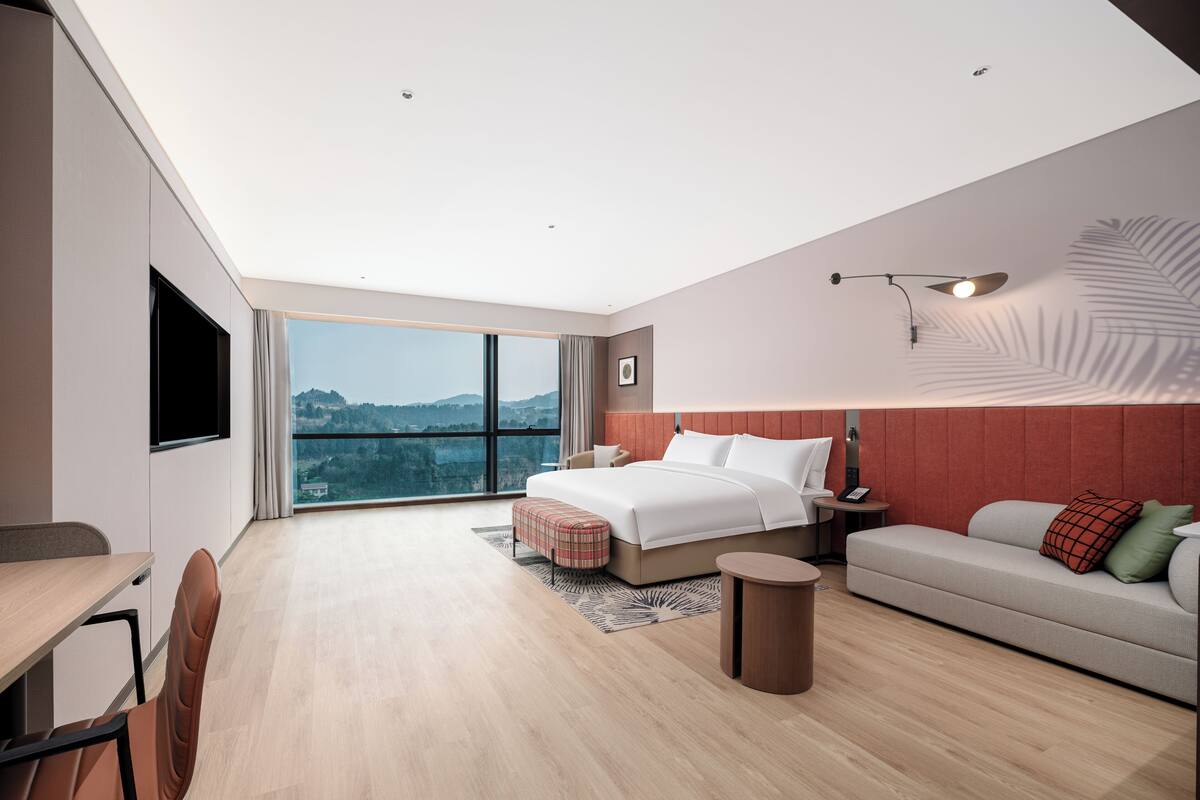 A modern hotel room with a large bed, a sofa, a desk, and a view of the city.