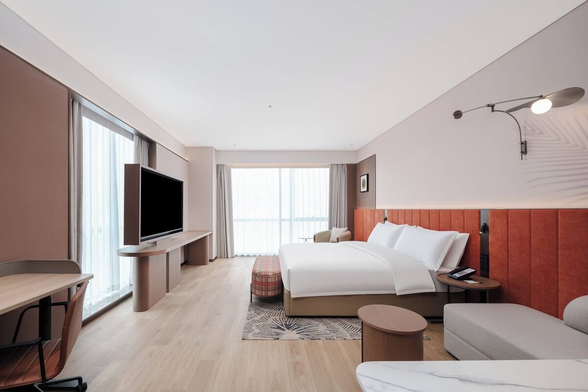 A modern hotel room with a large bed, a TV, a desk, and a seating area.