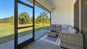 A modern outdoor seating area with a sofa and coffee table, a glass door leading to a grassy landscape, and a clear blue sky. - Palm Bay Luxury Haven (Palm Bay)