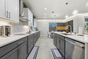 A modern kitchen with grey cabinets, white countertops, and stainless steel appliances. There is a dining area with a table and chairs, and a window with blinds. - Palm Bay Luxury Haven (Palm Bay)