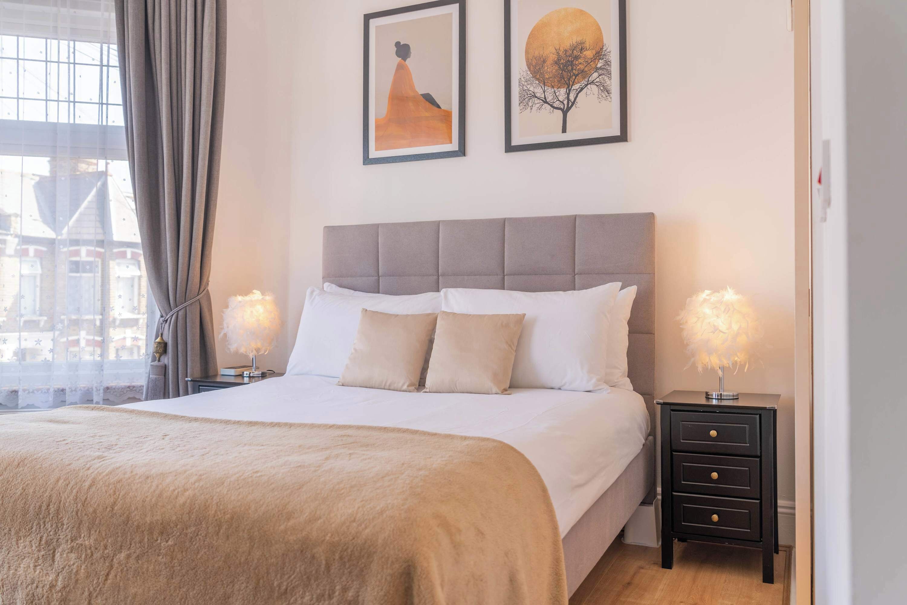 A hotel room with a large bed, two bedside tables, and a window with curtains. There are two framed artworks on the wall.