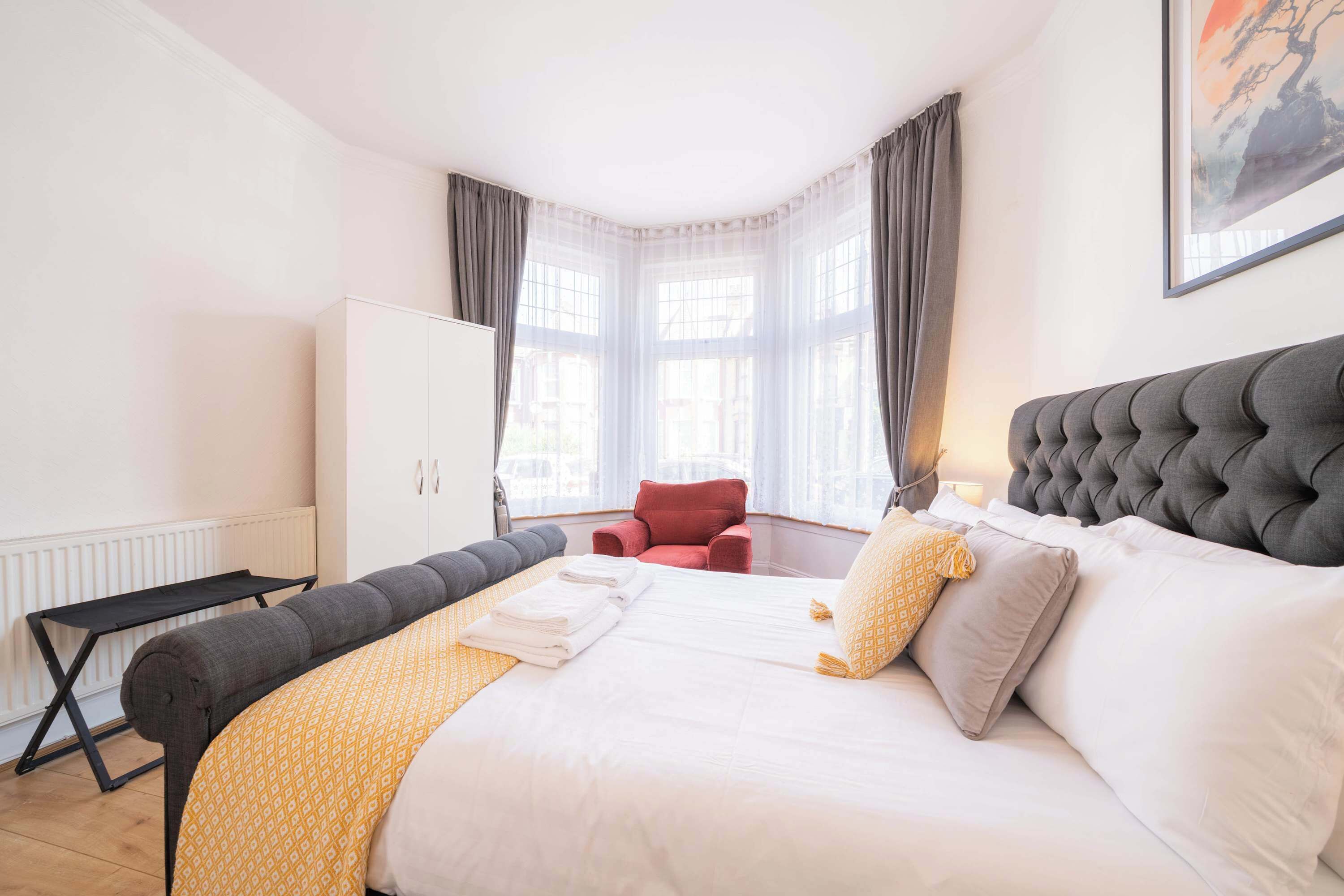 Une chambre d’hôtel avec un lit, une chaise rouge, une armoire blanche et une fenêtre avec des rideaux.