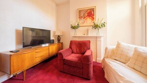 Una habitación de hotel con un aparador de madera, un sillón rojo, una cama con ropa blanca, un televisor de pantalla plana, un jarrón con flores y un cuadro en la pared.